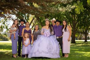 Outdoor quinceañera group photo with party wearing purple and lavender attire under tree canopy.