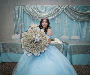 Quinceañera celebration with person in light blue ball gown holding large decorative money fan.