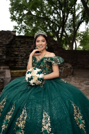 Elegant emerald green quinceañera ball gown with gold embroidery and white floral bouquet against stone wall backdrop.