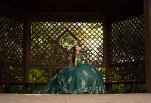 A person poses in an ornate teal ballgown inside a wooden gazebo with latticed walls.
