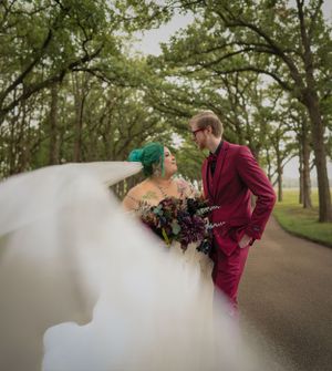 Romantic wedding photo featuring couple in burgundy suit and white dress with teal hair accessories on a tree-lined path.