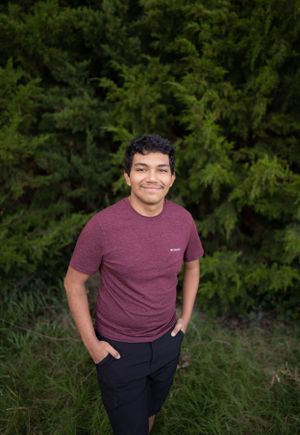 A person in a burgundy t-shirt poses casually against a background of evergreen trees.