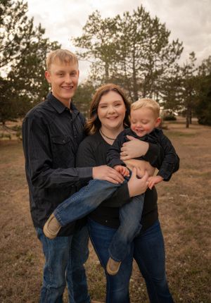 Family portrait of three people in casual clothing outdoors with pine trees.