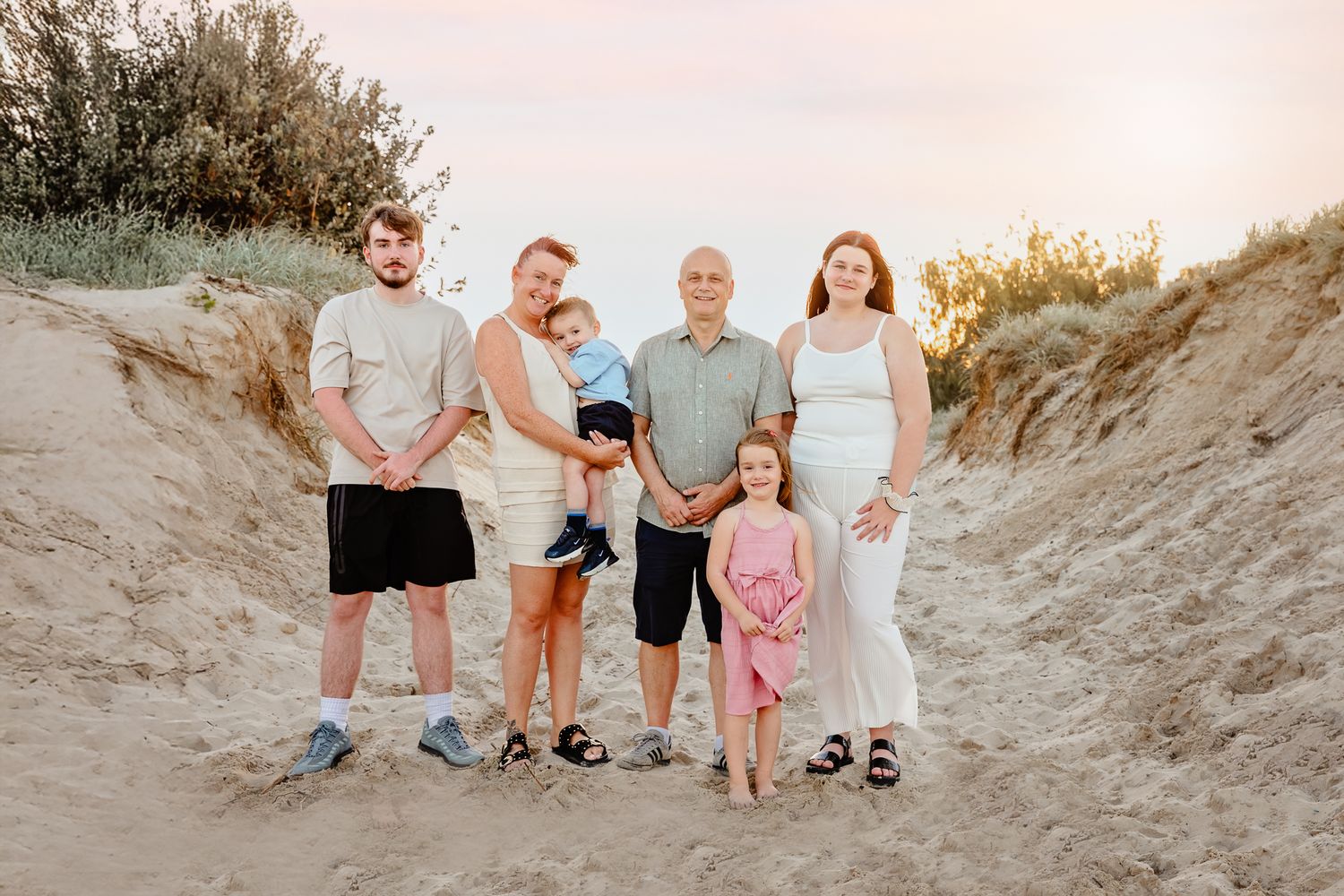 Family of six posing on sandy beach dunes at sunset, including pregnant woman and young children.