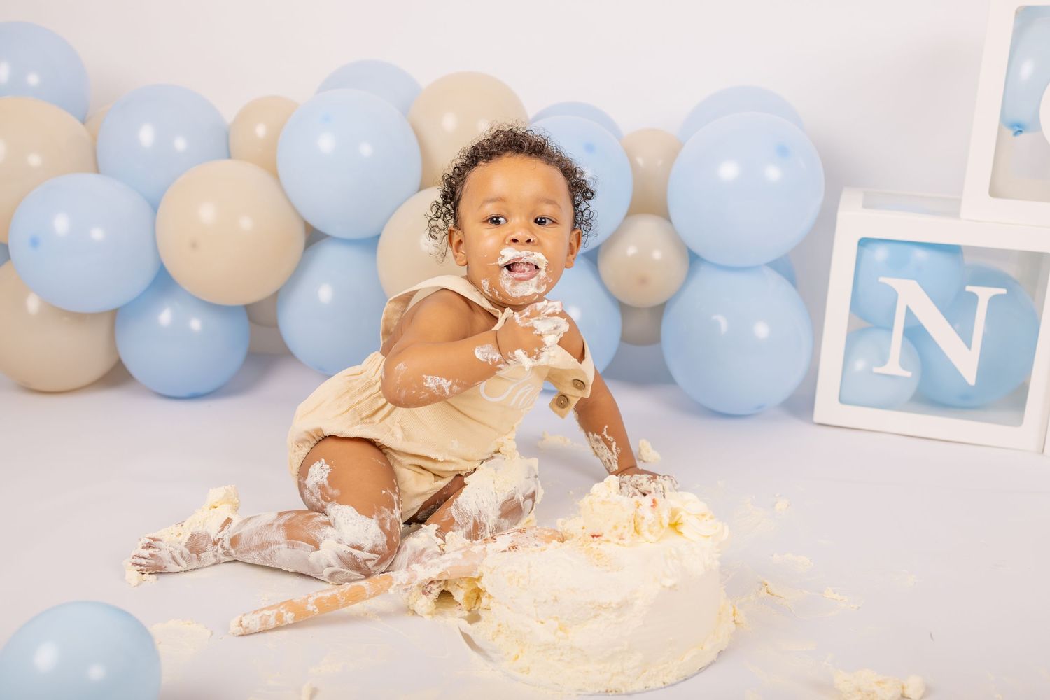 Baby in white outfit sits on floor surrounded by blue and cream balloons during cake smash photo session.
