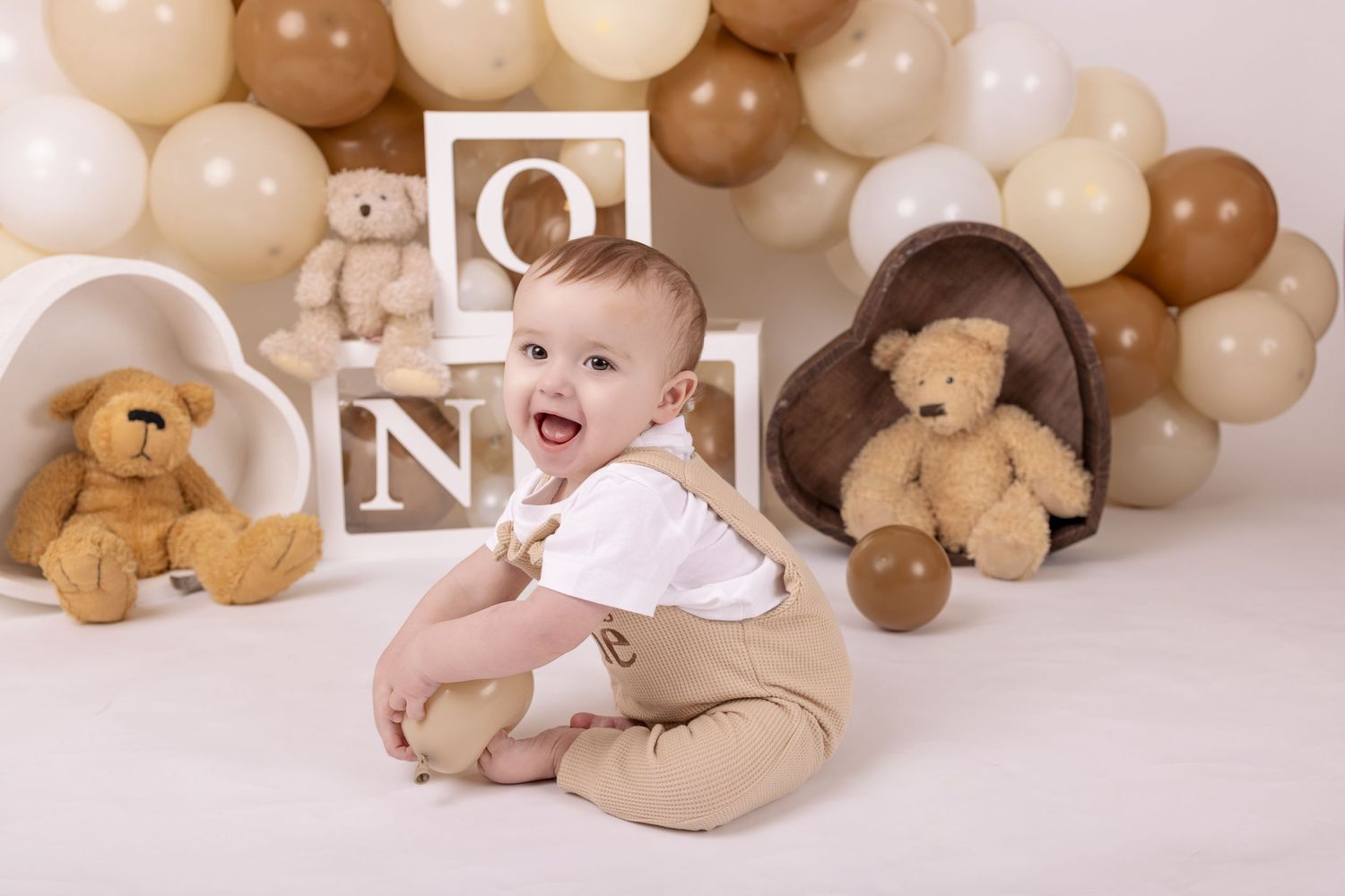 A baby photo collage with brown and beige balloons, teddy bears, and soft pink accents creates a warm nursery setting.