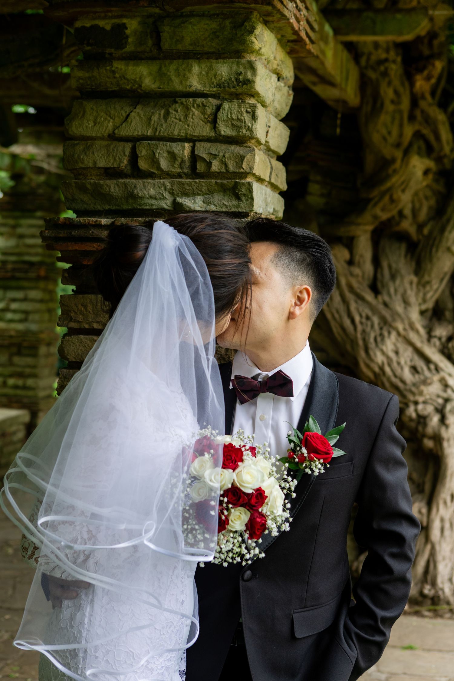 A romantic wedding couple shares intimate moments in a garden setting with red and white floral bouquet details.