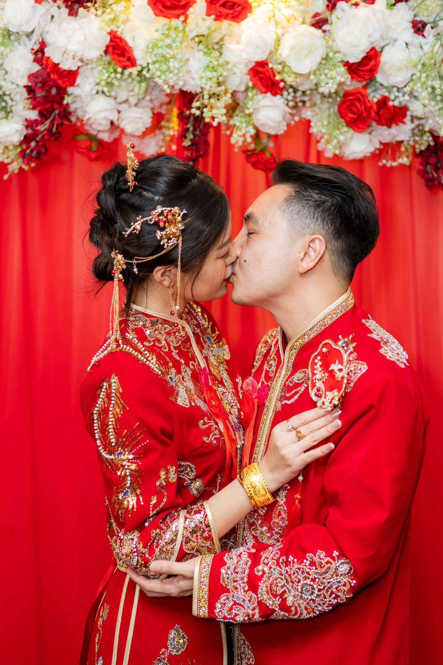 Traditional Chinese wedding couple in red qipao dress and suit share romantic kisses against floral backdrop.