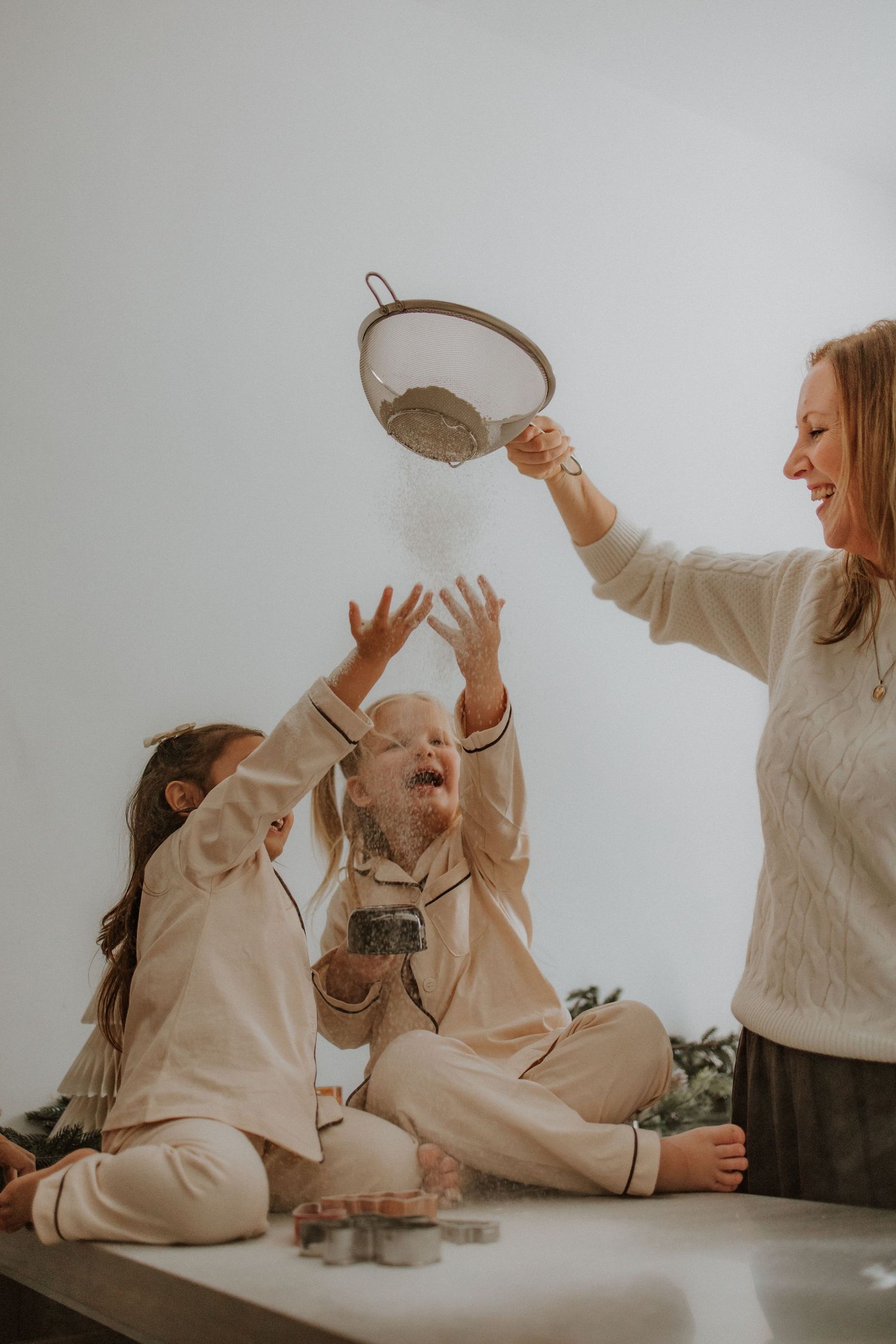 A smiling mother sifts flour over two laughing children in matching cream pajamas during a fun holiday baking session.