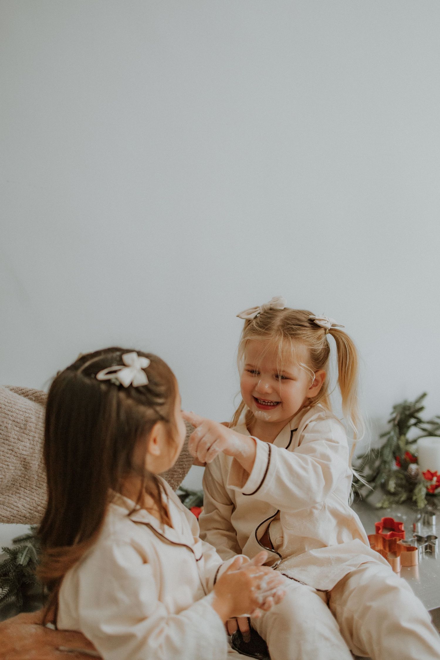 Two young girls in matching cream pajamas laugh and play together near Christmas greenery and decorations.