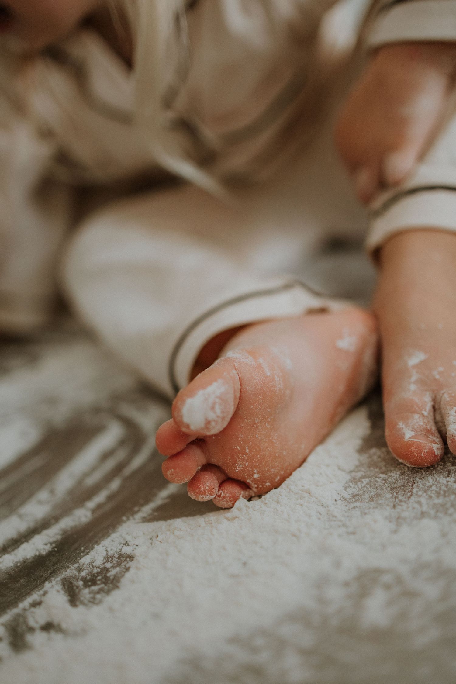 Close-up of a baby's flour-dusted bare foot on a white powder-covered surface, wearing a neutral-toned outfit.