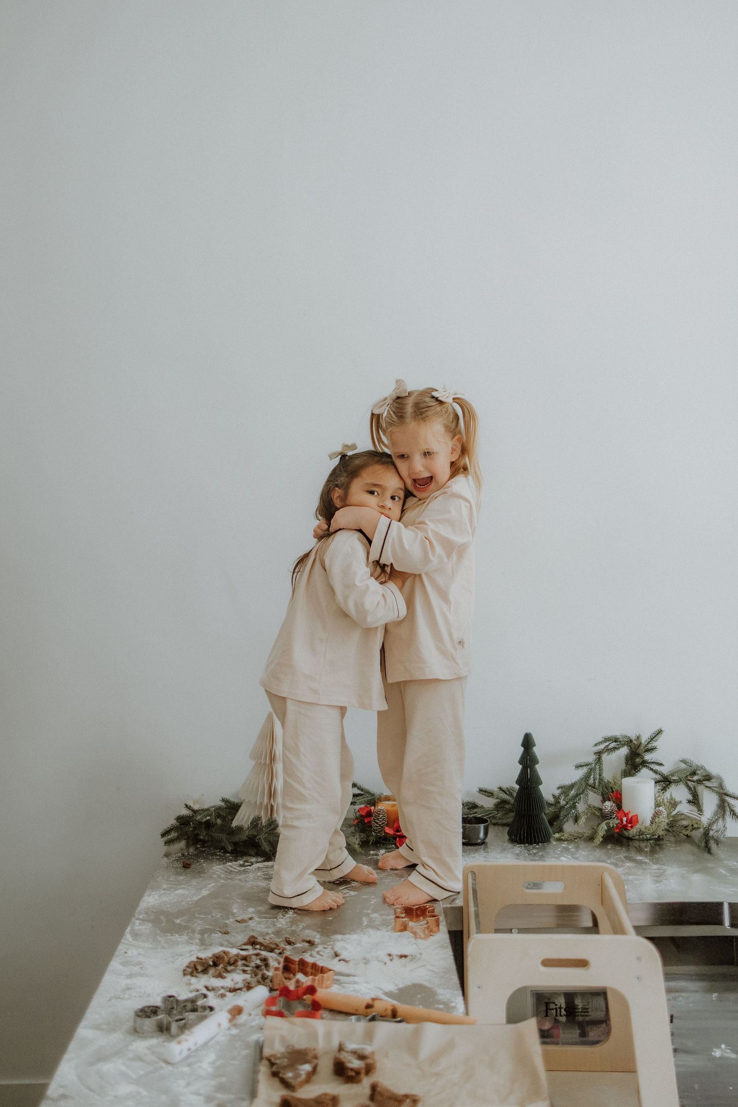 Two young girls in matching cream pajamas hugging on a table surrounded by Christmas decorations and cookie cutters.