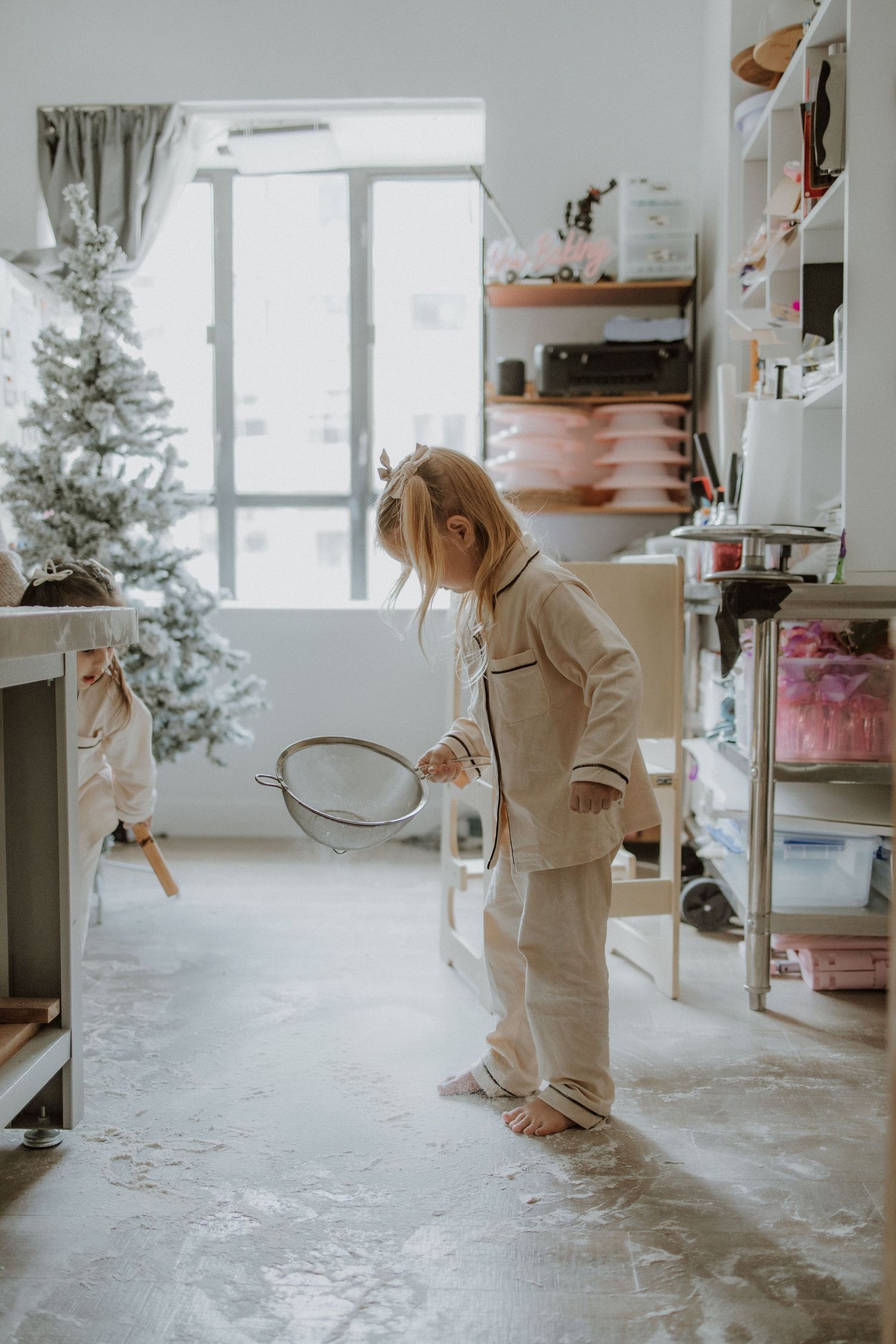 Young child in cream pajamas holding a strainer, playing indoors near a Christmas tree with flour on the floor.