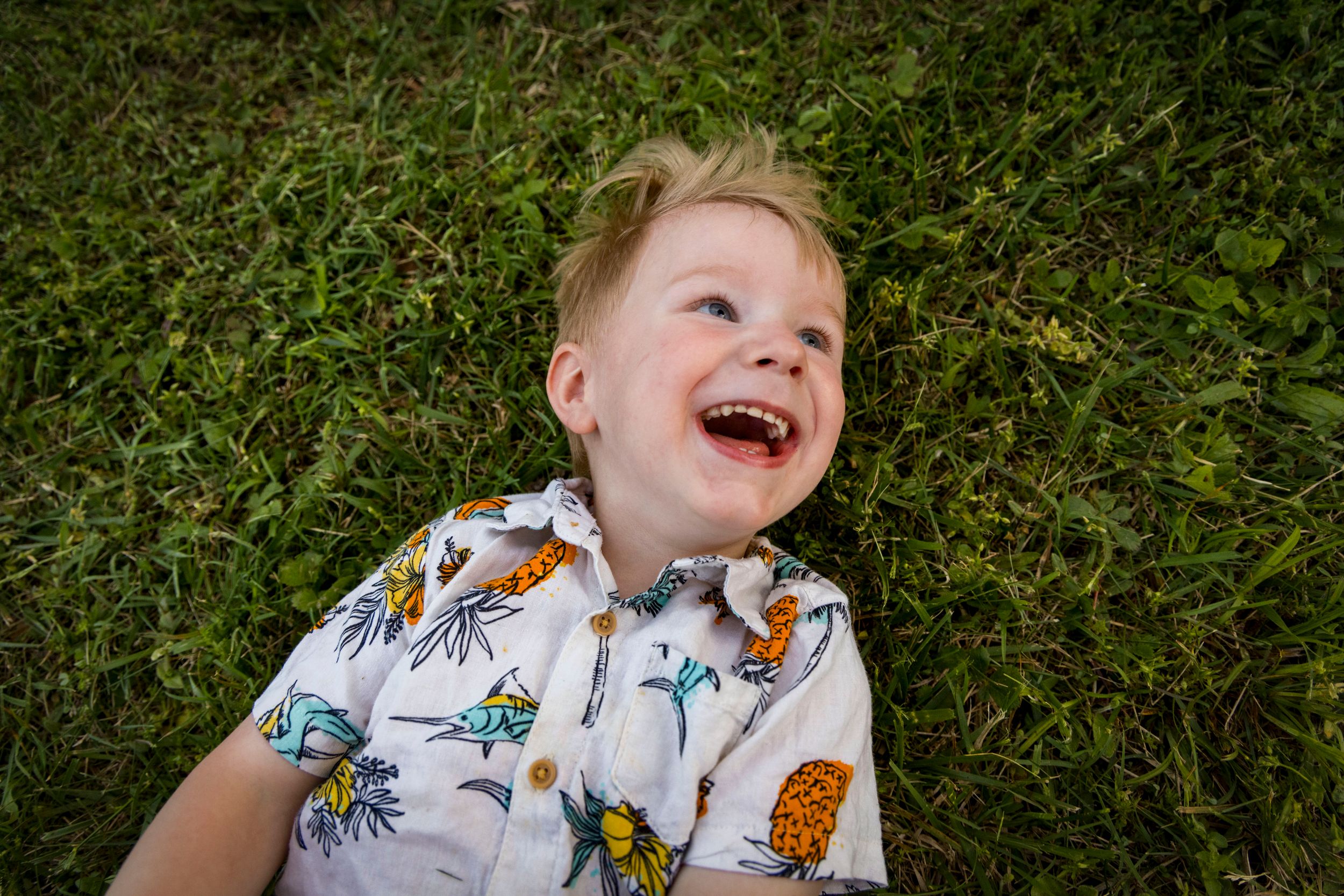 A joyful young boy laughing while lying on green grass, wearing a colorful tropical patterned shirt.