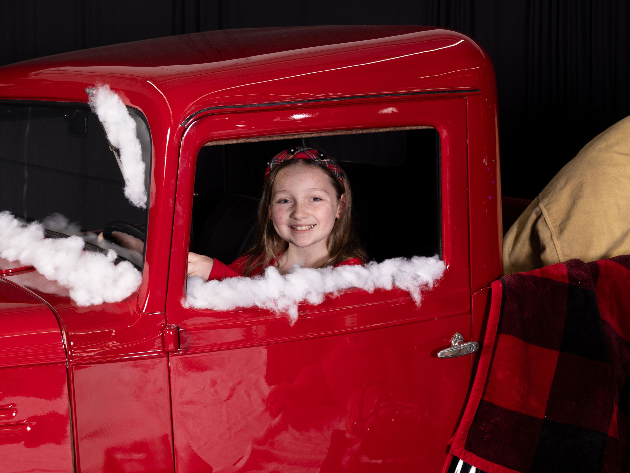 Young girl smiling inside a decorated red vintage truck with snow accents during a Christmas photo shoot.