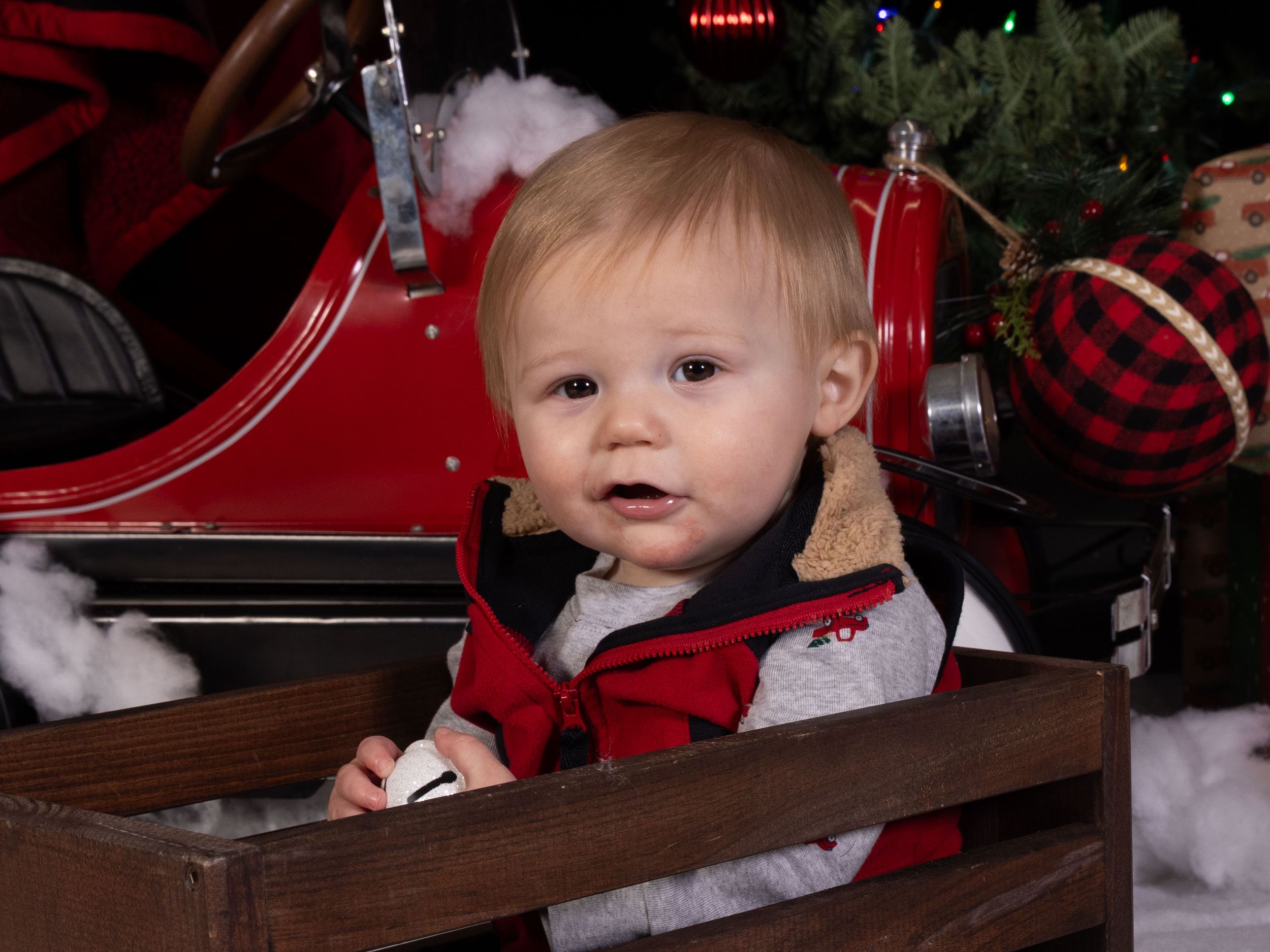A baby in a red plaid outfit sits in a wooden crate near a red vintage car and Christmas tree decorations.