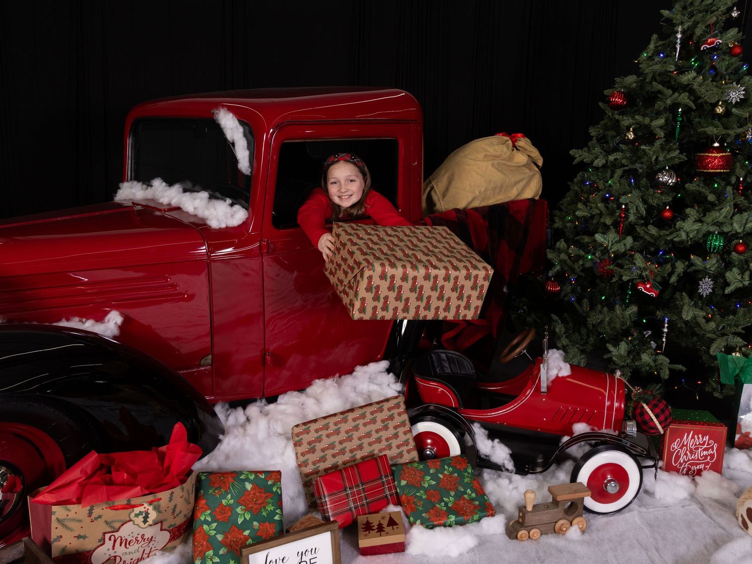 A red vintage toy truck sits in a Christmas holiday display with decorative trees and wrapped presents and girl leaning out of the window