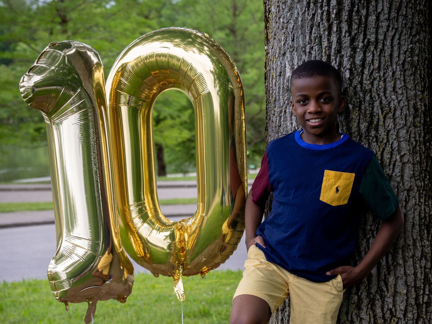 A large gold number 10 balloon decoration stands next to a young boy smiling while leaning on a tree trunk in an outdoor park setting