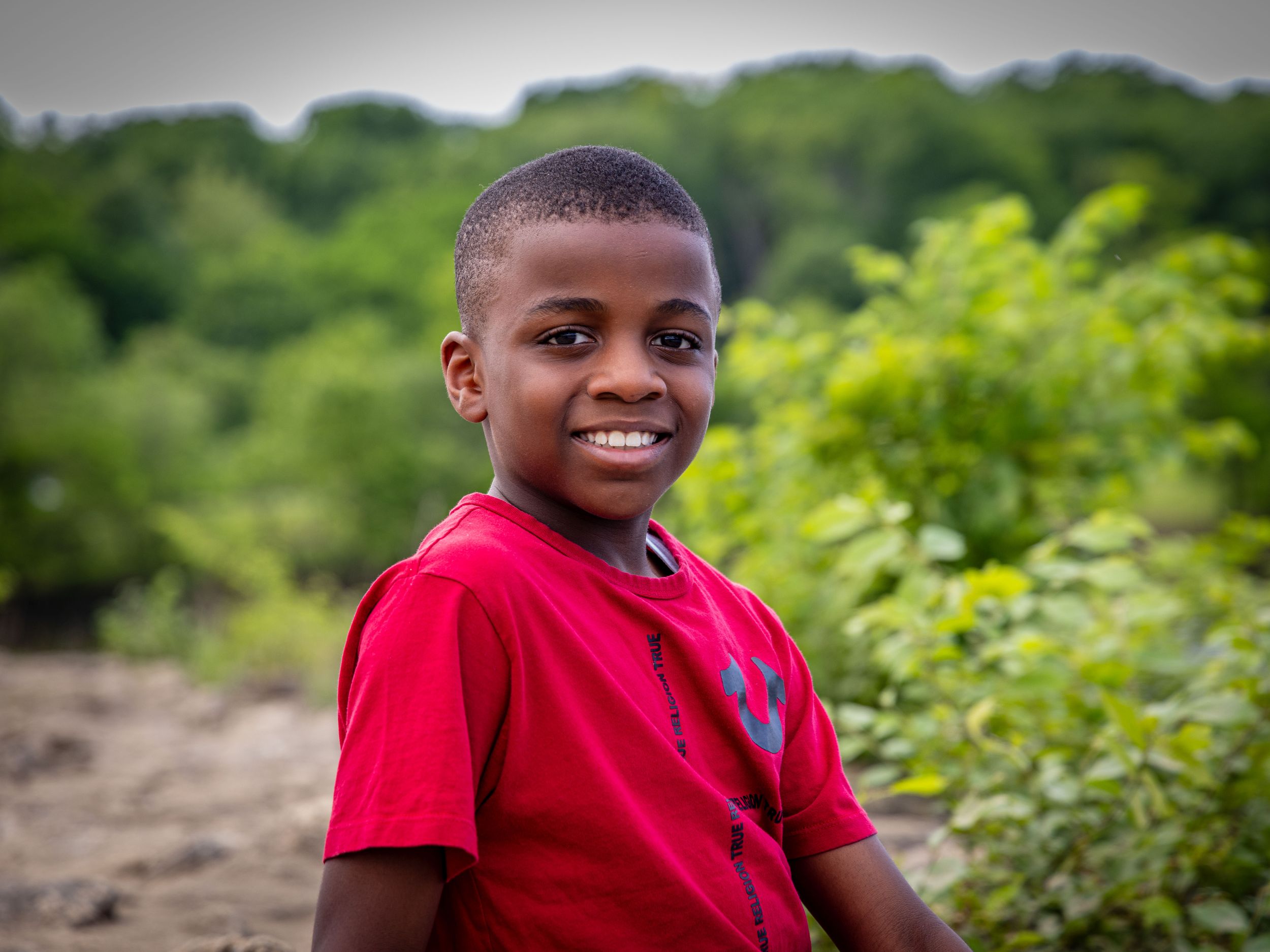 A young boy in a red shirt smiling outdoors in a lush green environment.