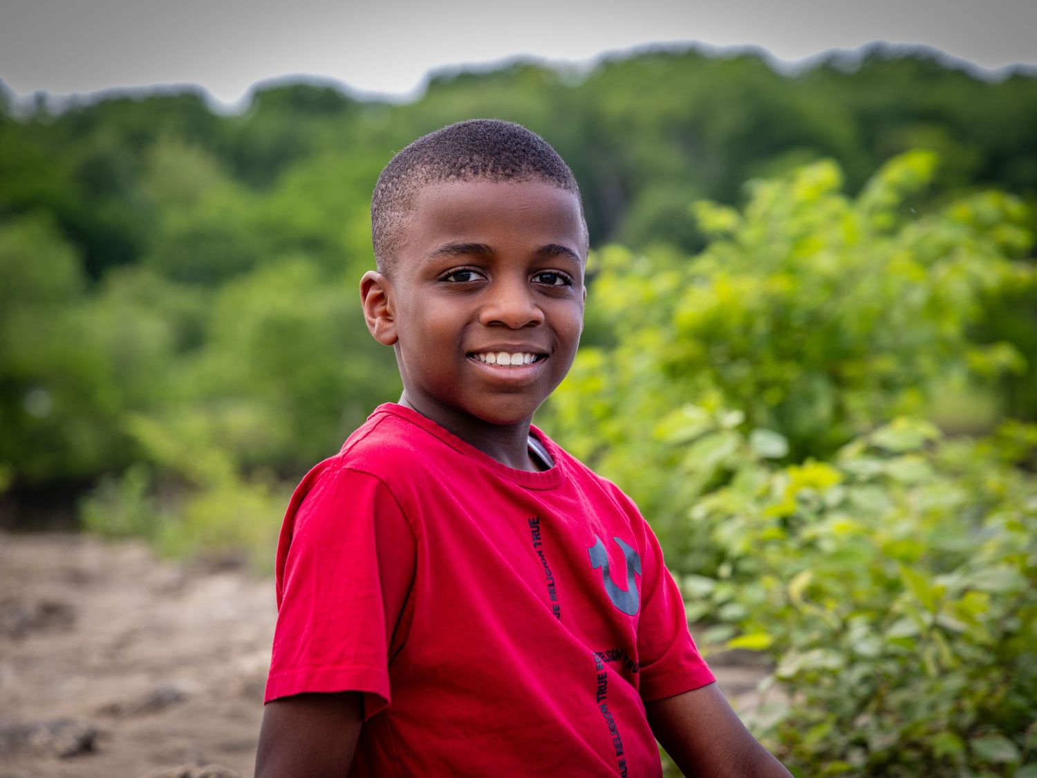A young boy in a red shirt smiling outdoors in a lush green environment.