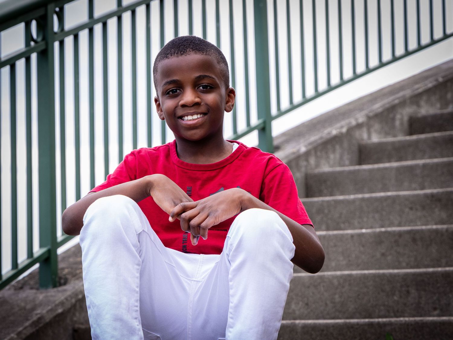 A young boy in a red shirt and white pants smiles while sitting on concrete steps outdoors.