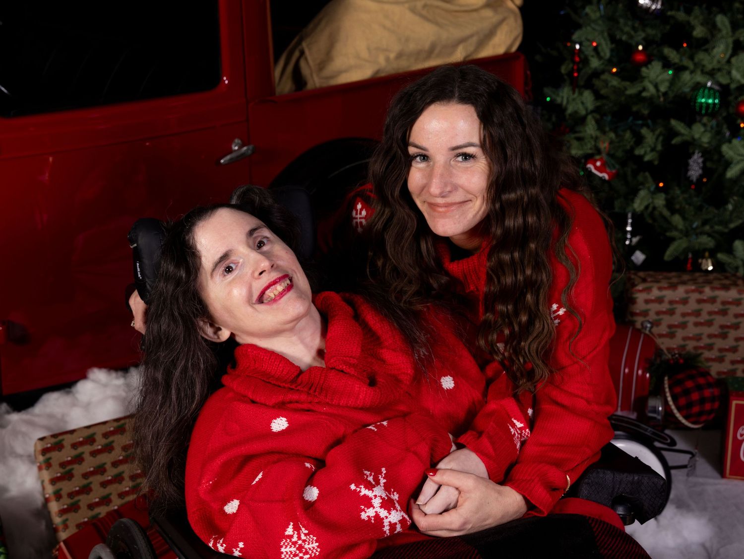 Sisters in festive red sweaters posing playfully in front of a decorated Christmas tree and vintage truck