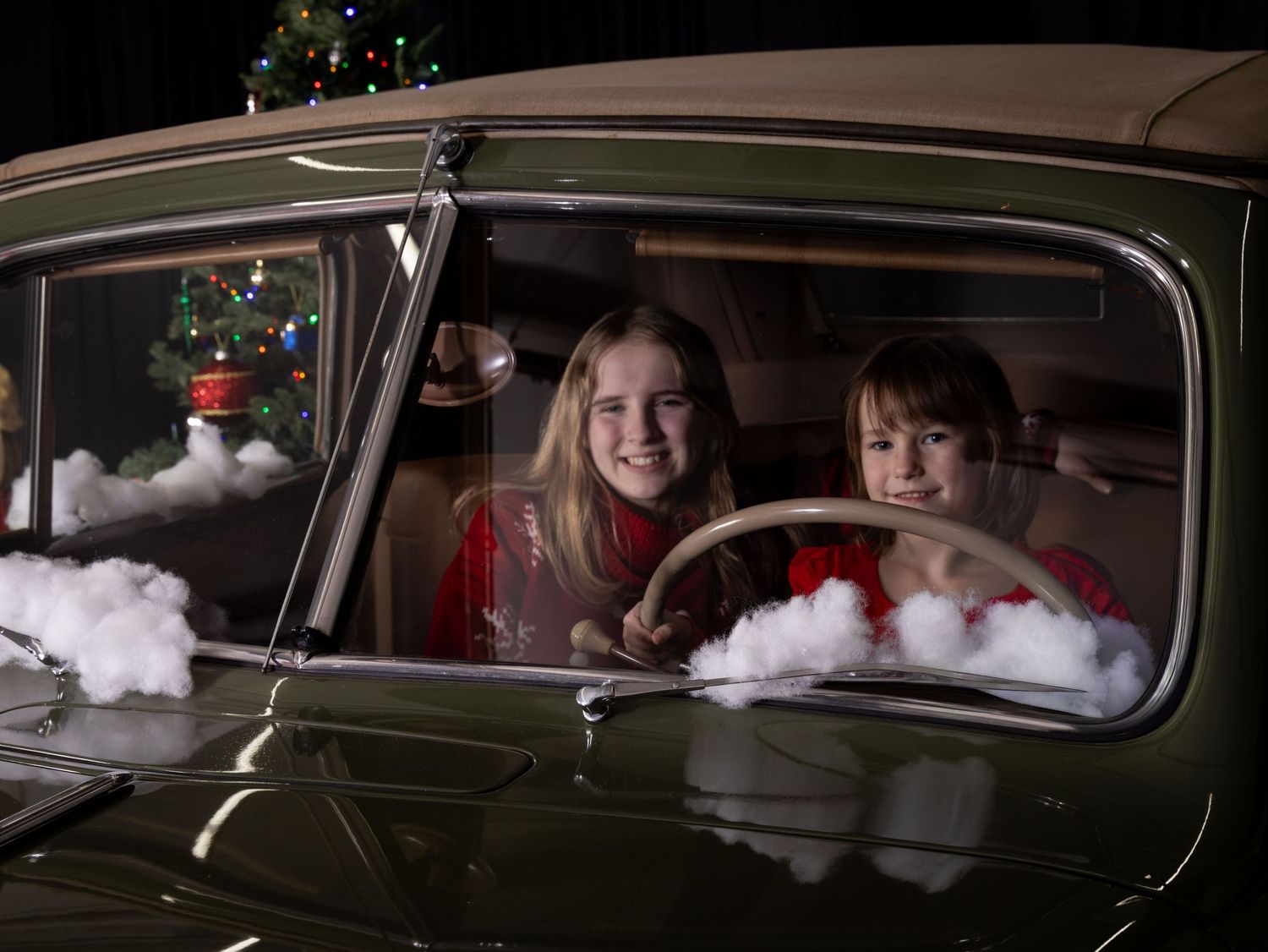 Children sit in a vintage car with Christmas decorations and snow visible through the windows.