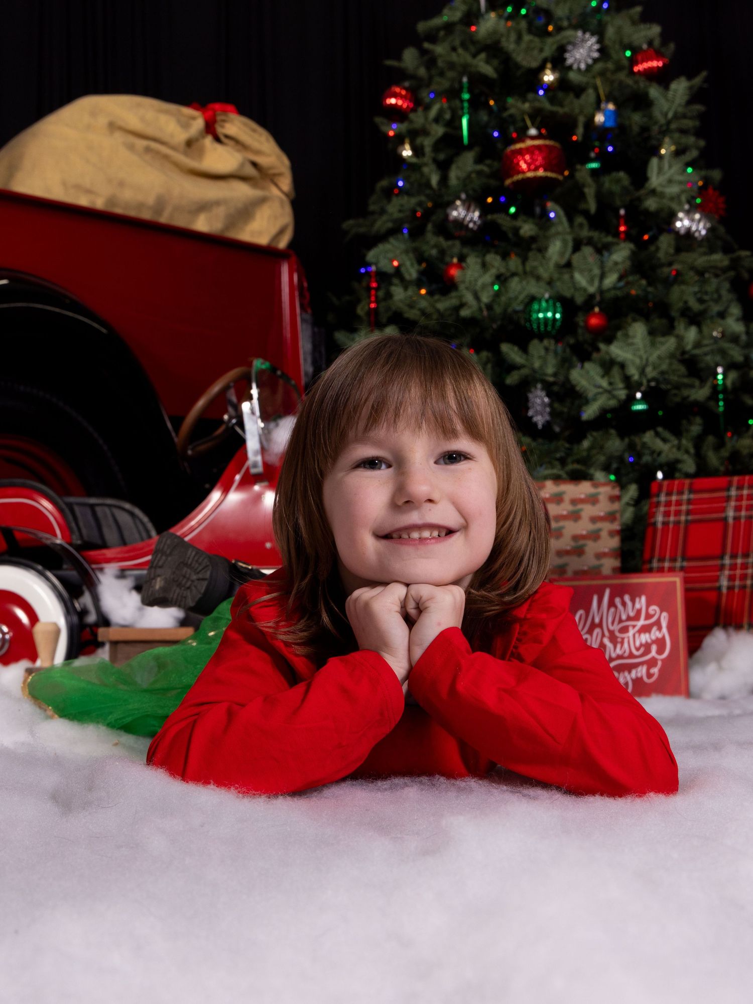A Christmas photo showing a child lying on white snow with red decorations and a Christmas tree in the background.