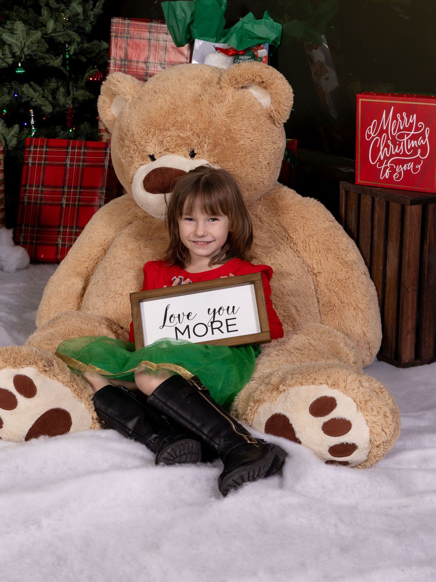A holiday photo showing a child sitting with a large teddy bear holding a Love you more sign in a snowy Christmas setting.