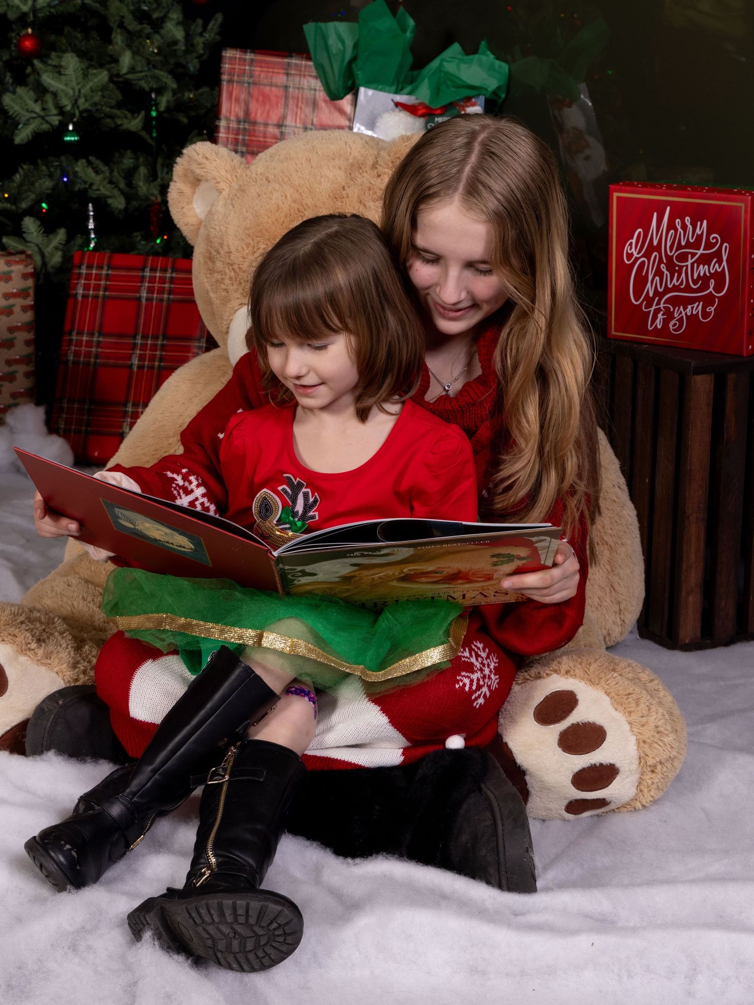 Sisters reading a book while sitting on plush teddy bear surrounded by Christmas decor in a festive holiday setting.