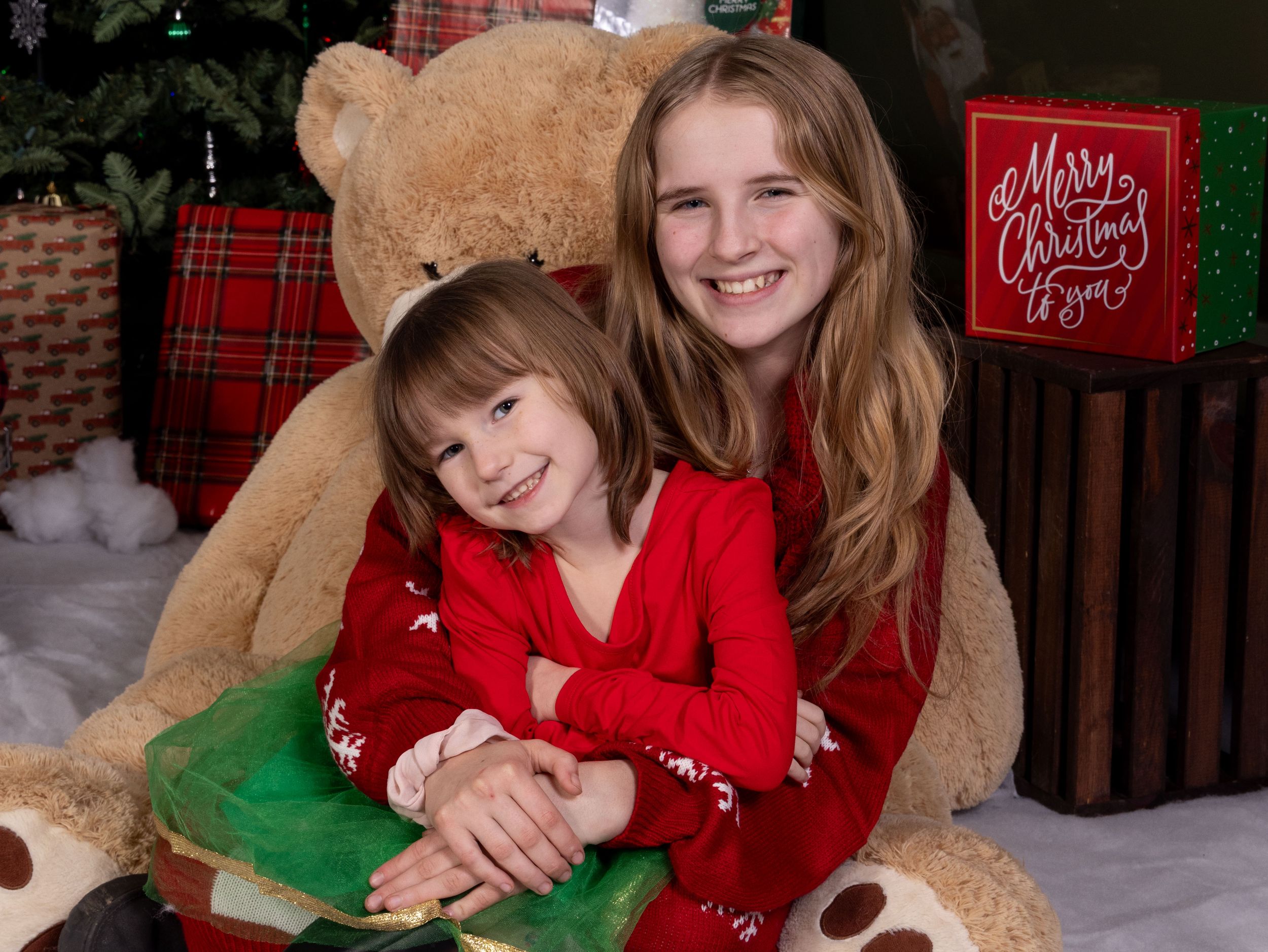 Two girls in red Christmas outfits smile with a large teddy bear and festive gifts near a decorated tree.