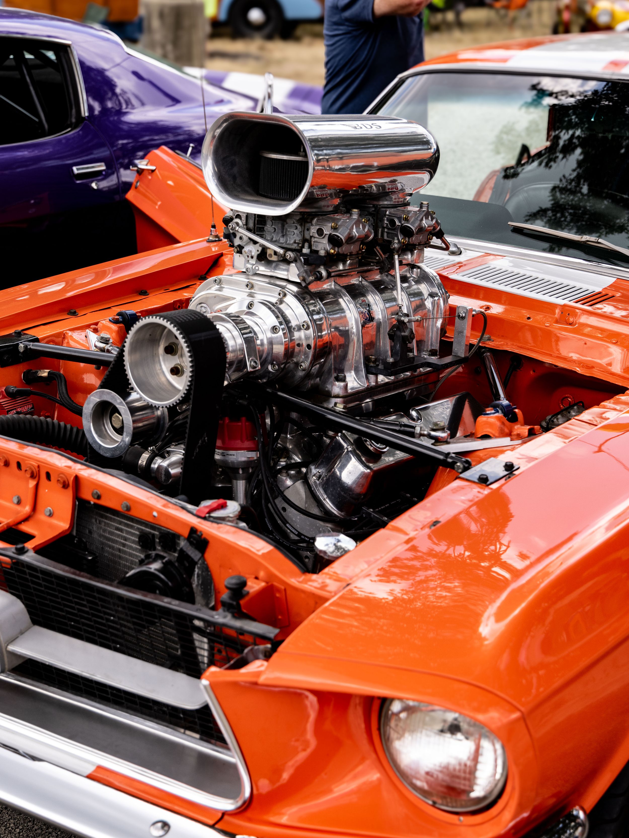 Custom orange muscle car with massive supercharged V8 engine and chrome blower scoop at a car show.