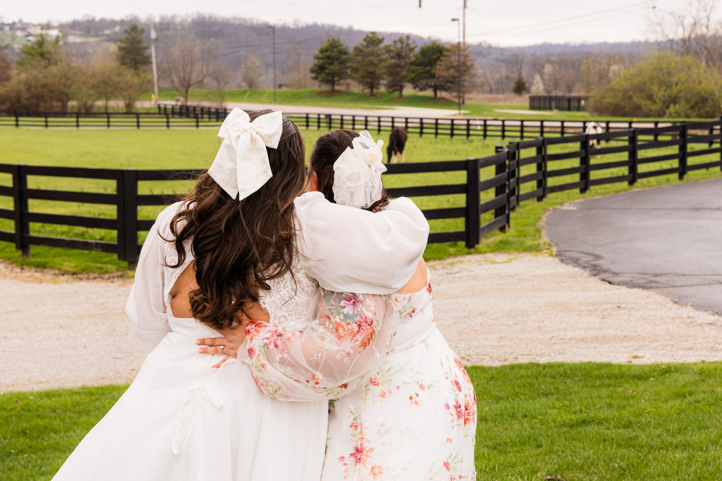 Two girls in white dresses with large bows hugging outdoors near a black fence on a green farm.