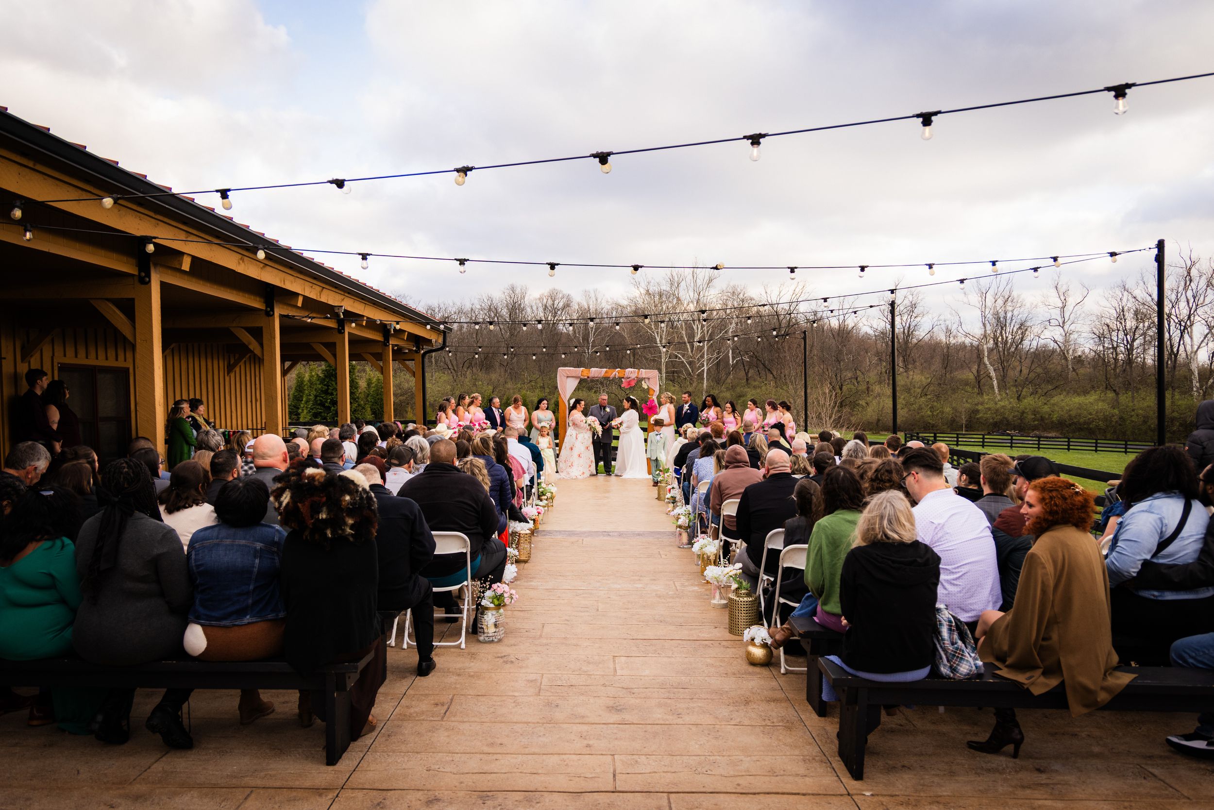 Outdoor wedding ceremony on a brick patio with string lights, guests seated in rows, and couple at floral arch altar.