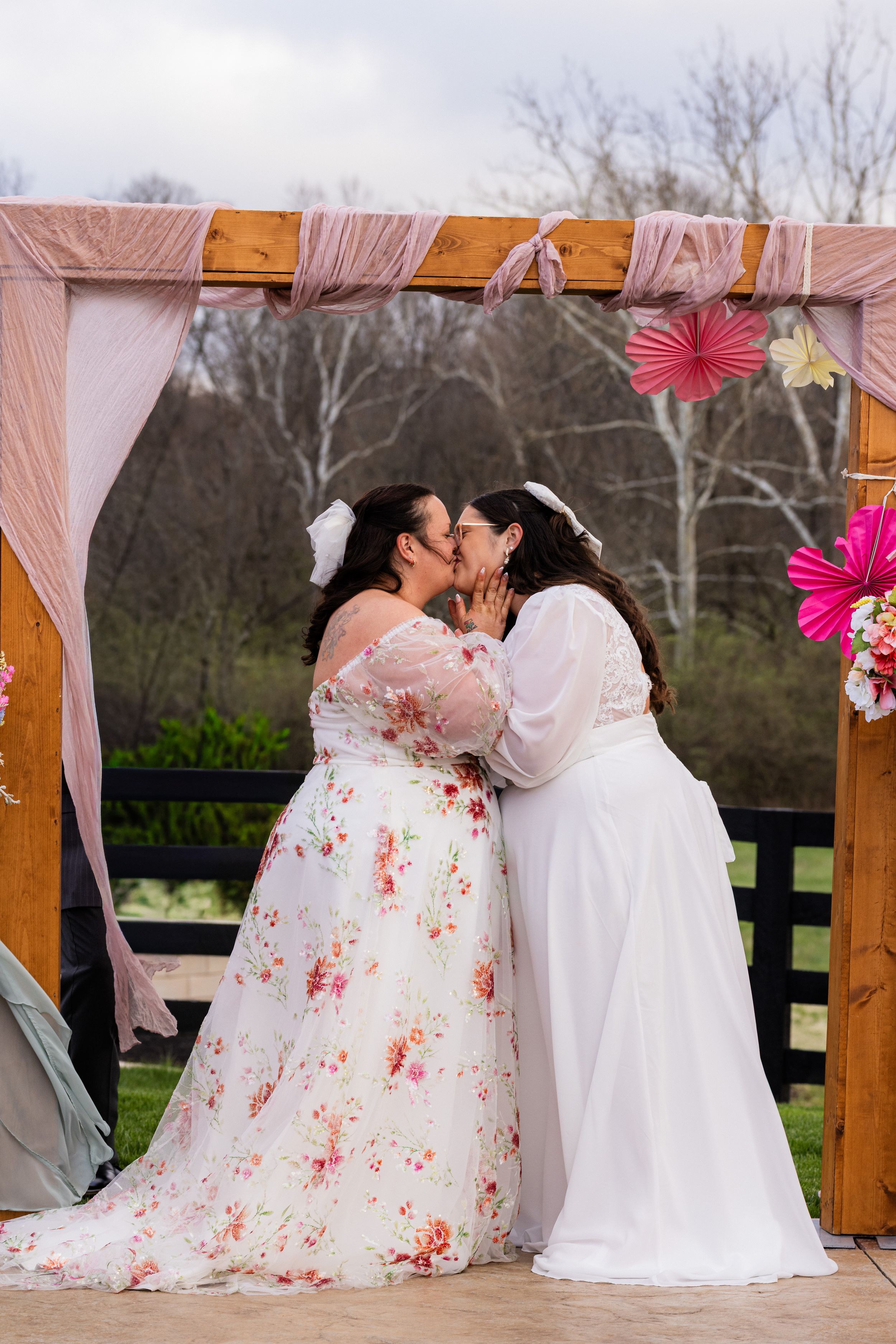 Two brides share their first kiss at an outdoor wedding ceremony beneath a decorated wooden arch with pink draping.