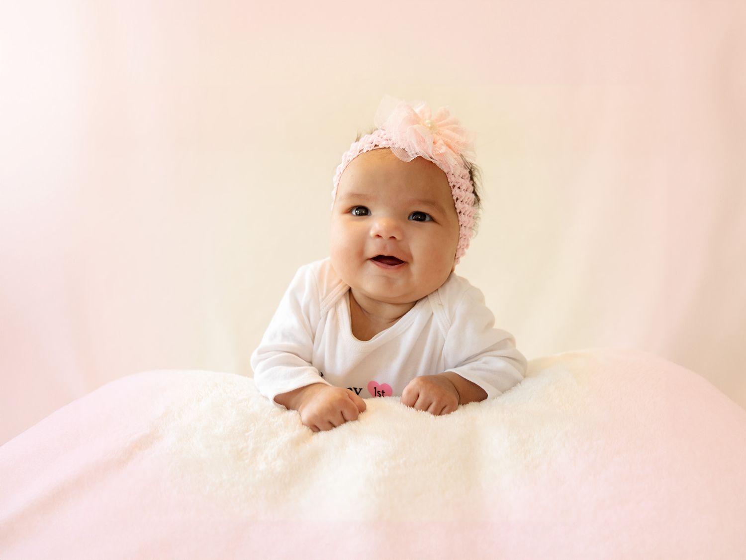 Baby in white outfit lying on soft white blanket with lace headband and cute laughter smile.