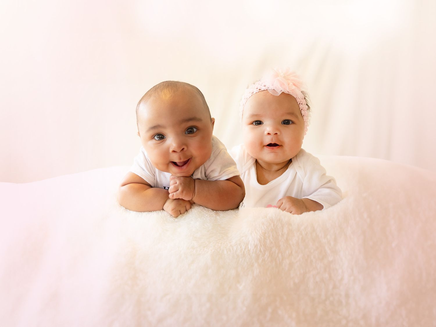 Two babies in white outfits lying together on soft white blanket, both smiling.