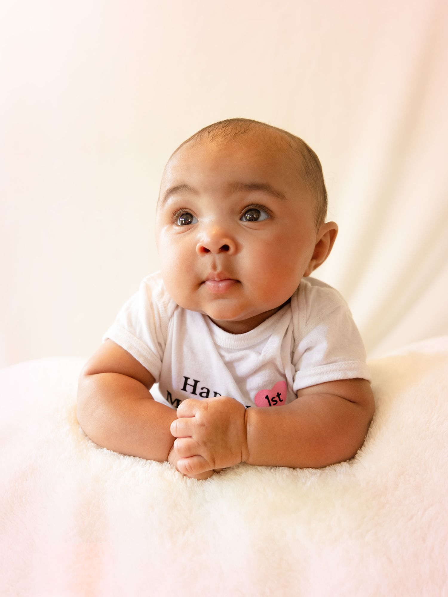 A baby in a white onesie rests on a soft white blanket while looking upward with a gentle expression.