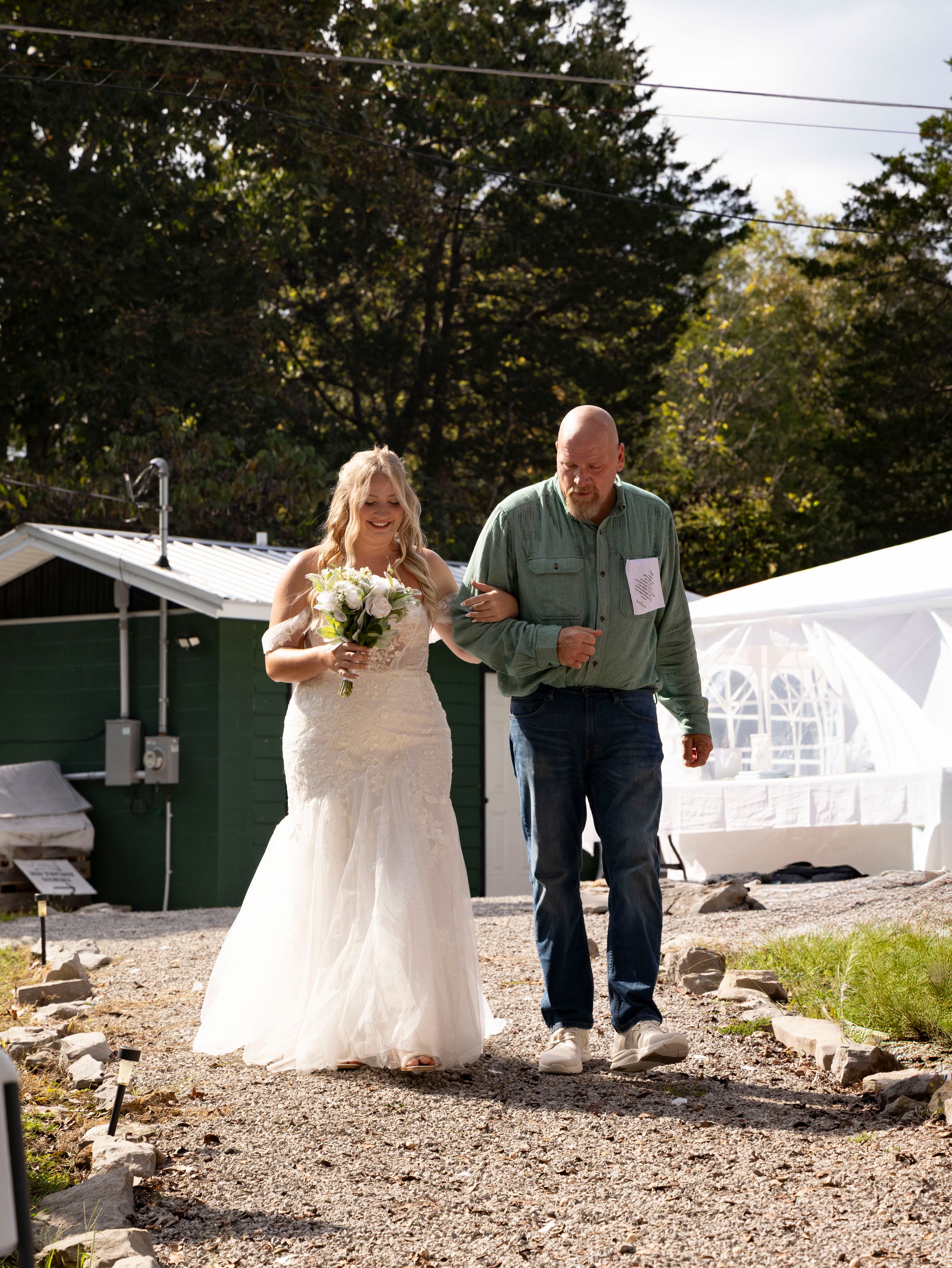 Bride in white lace gown holding bouquet walks with man in green shirt and jeans on gravel path at outdoor wedding.