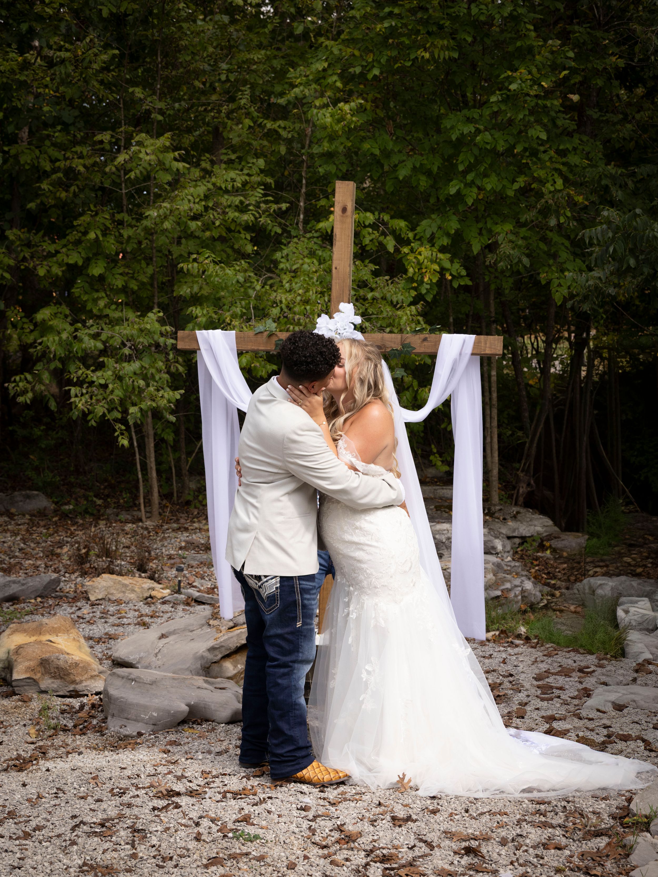 Bride and groom share first kiss at outdoor wedding ceremony with rustic wooden cross altar draped in white fabric.