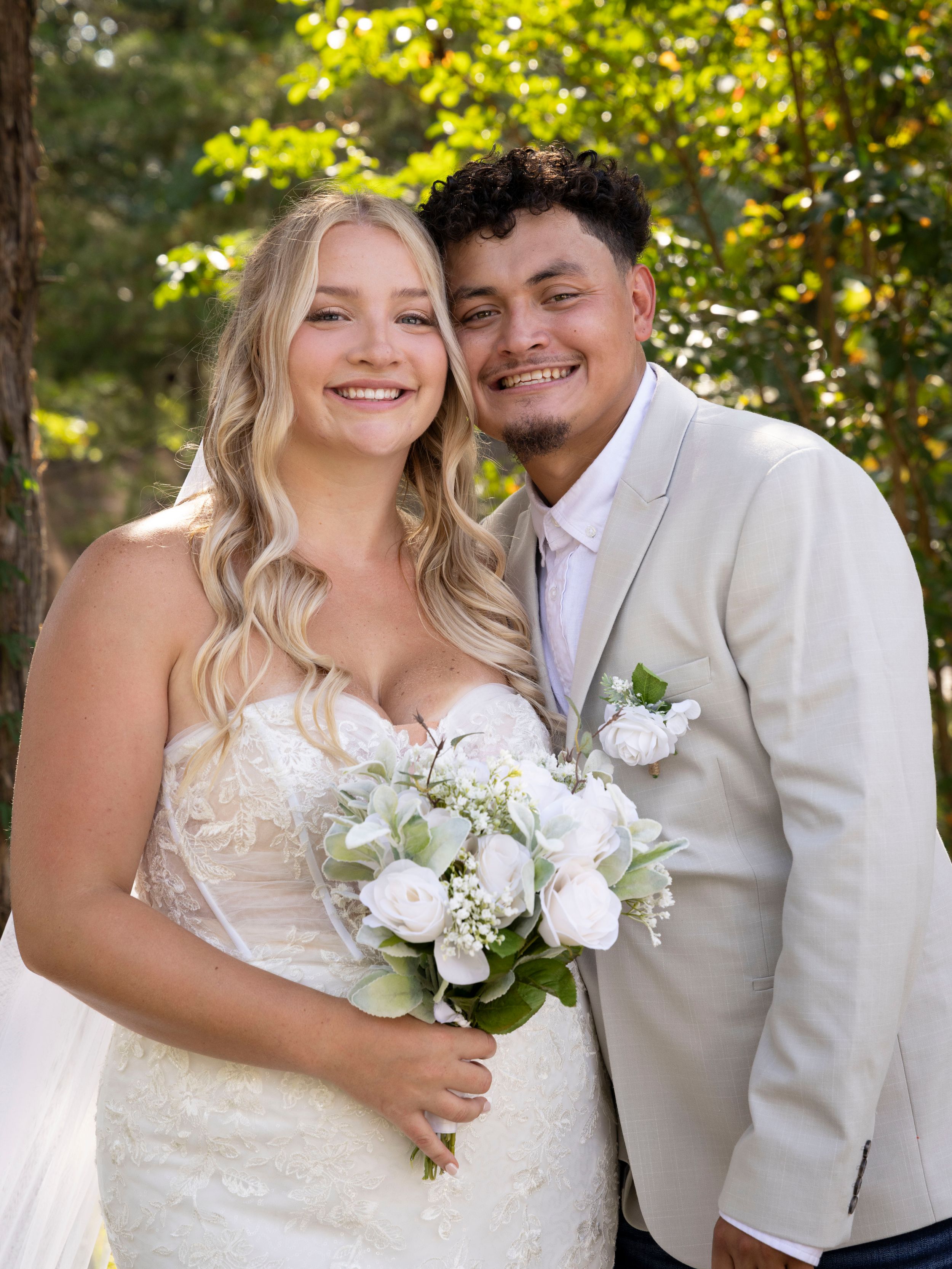 A smiling bride in a white lace gown holds a bouquet beside a groom in a tan suit outdoors.