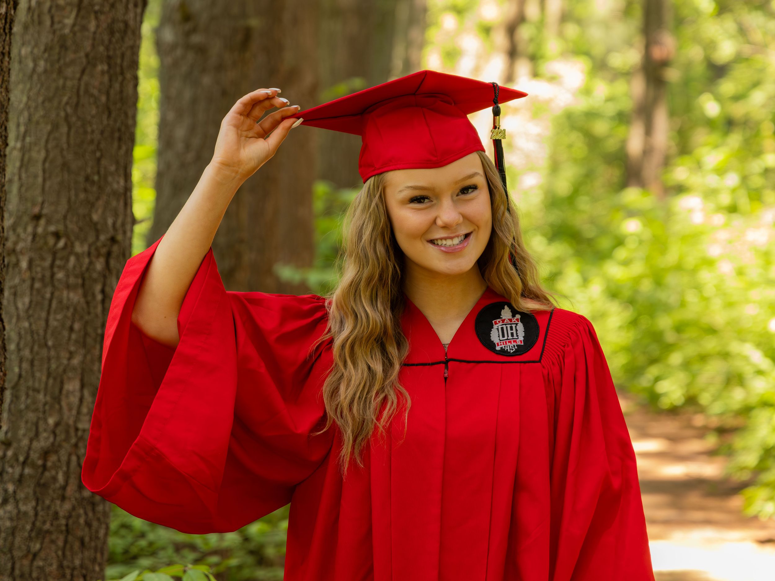 Graduate in bright red cap and gown smiles while posing for a photo outdoors in a wooded setting.