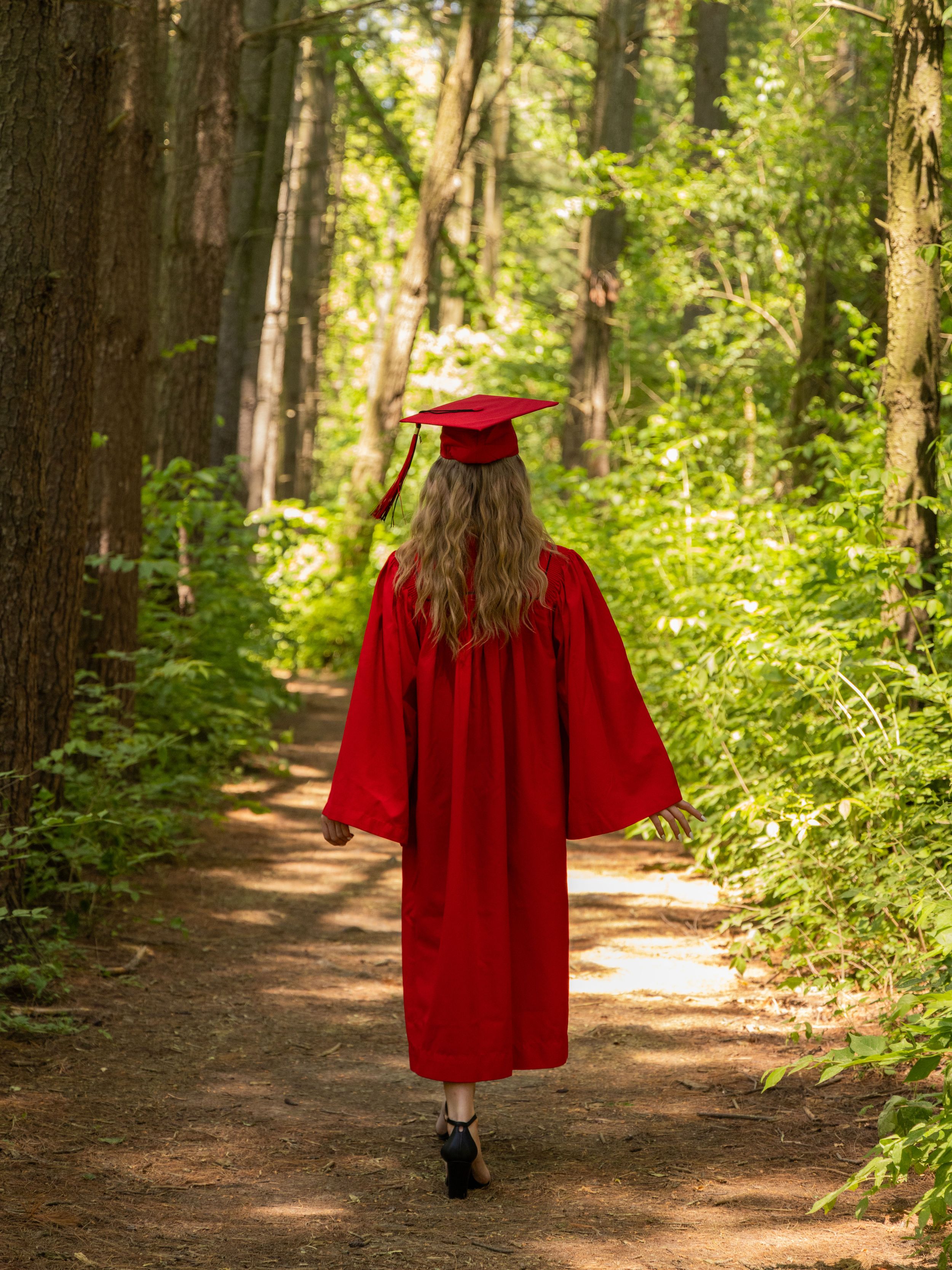 A graduate in red cap and gown walks along a sunlit forest path surrounded by lush green trees.