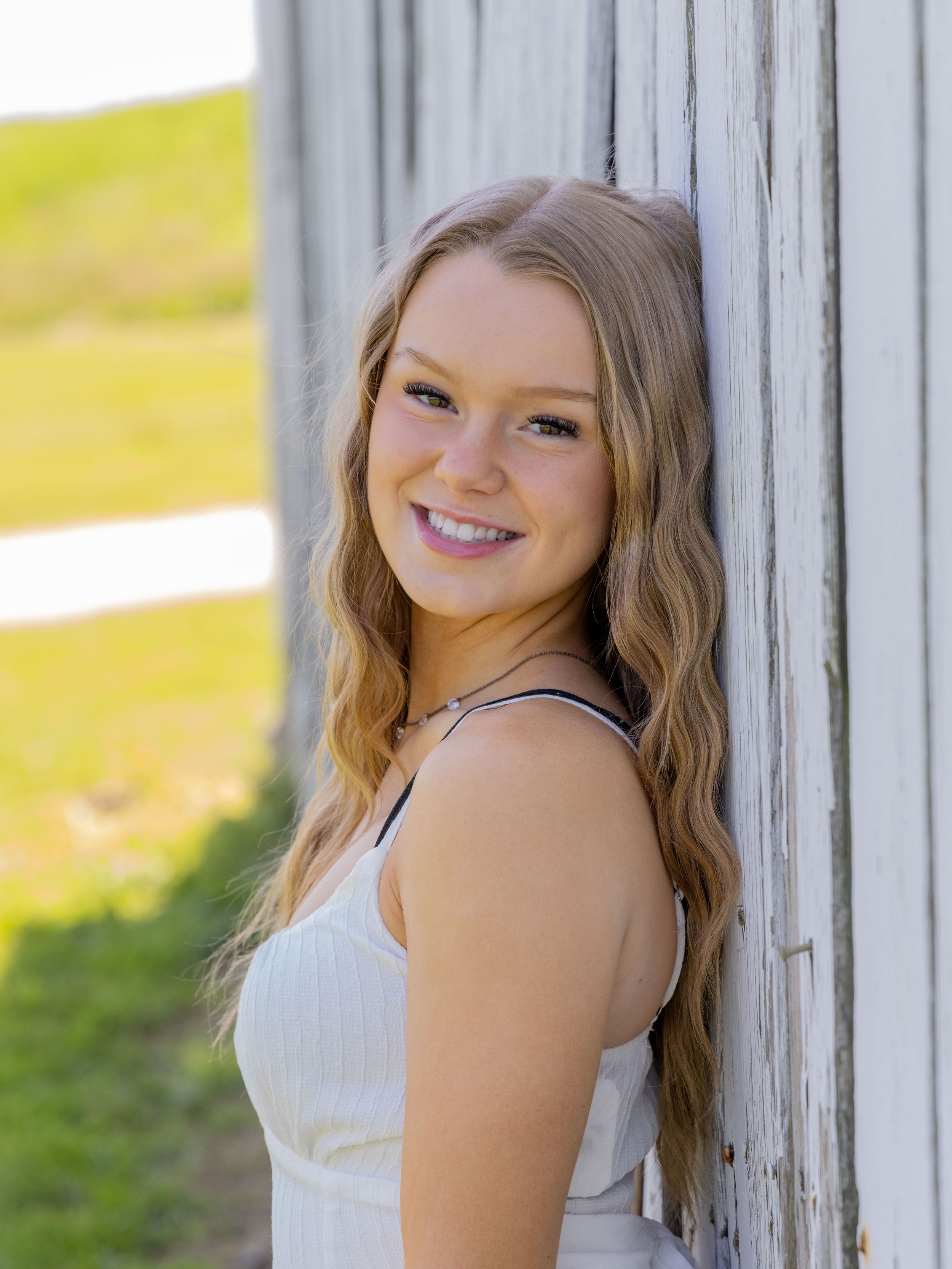 Young woman in white dress smiling while leaning against rustic white wooden wall with blurred green background.