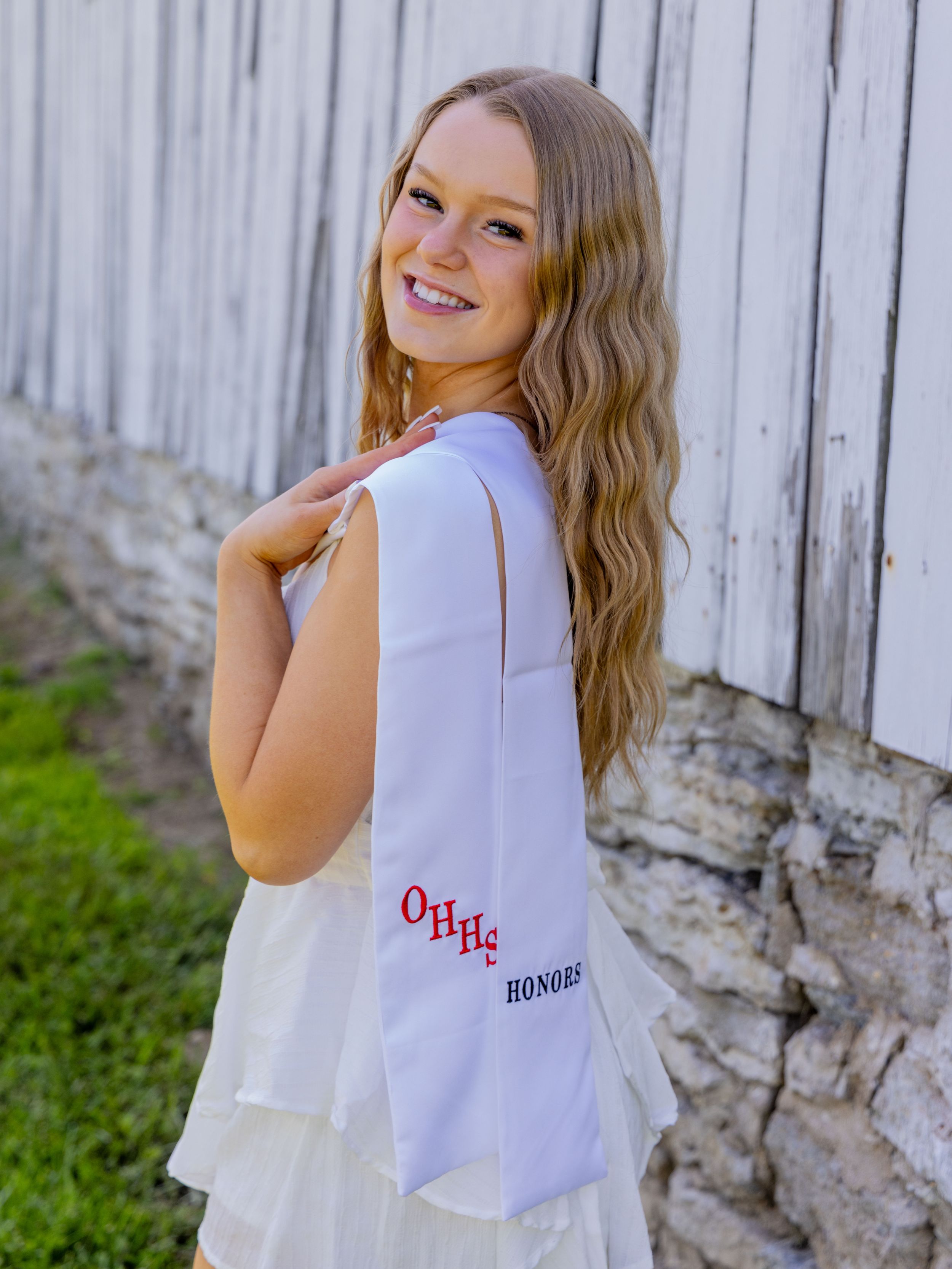 Girl in white dress smiles while posing against a white fence and stone wall holding highschool graduation shash embroidered with OHHS