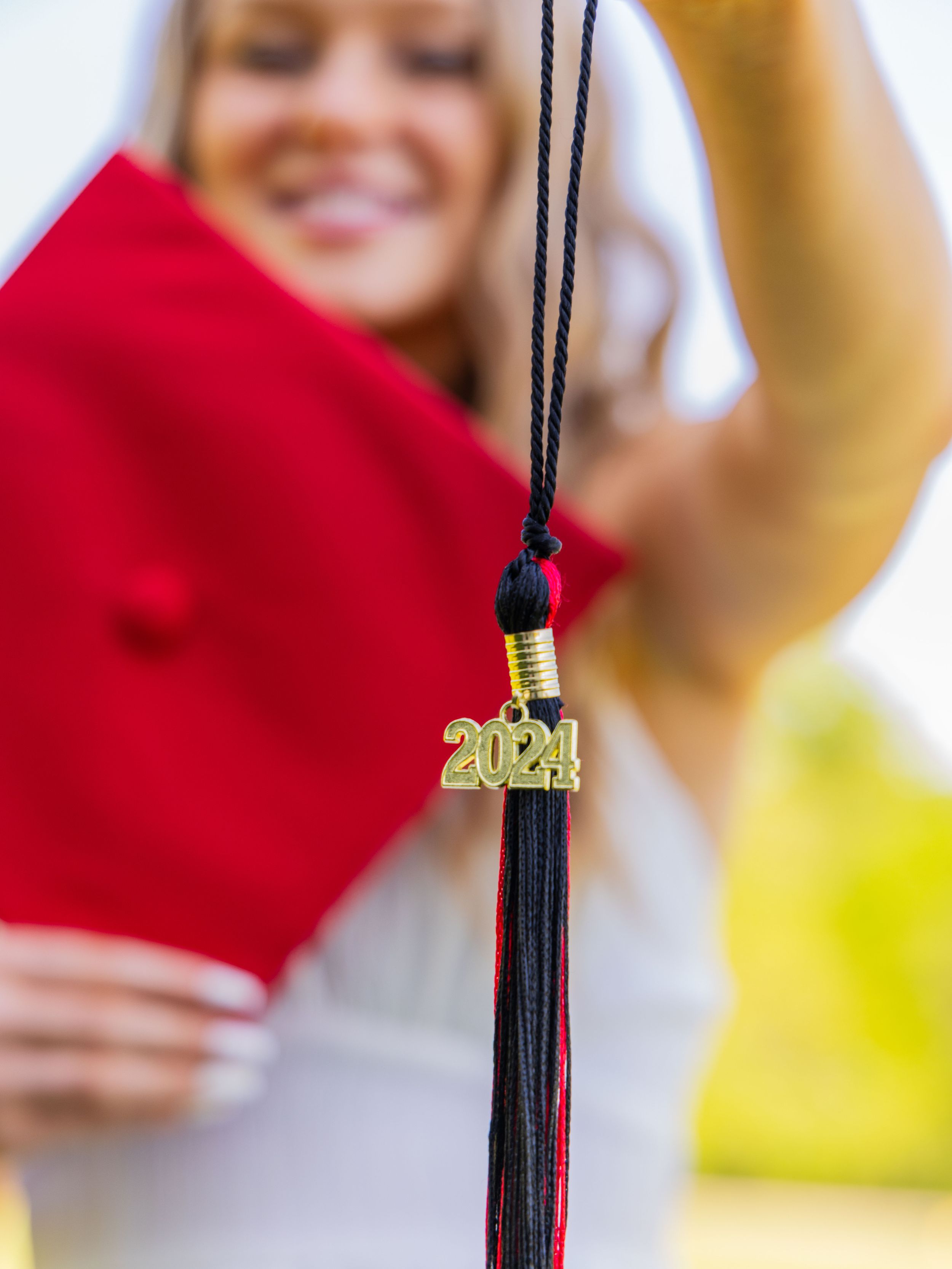 A black graduation tassel with gold 2024 charm hanging in focus with red graduate cap and blurred background.