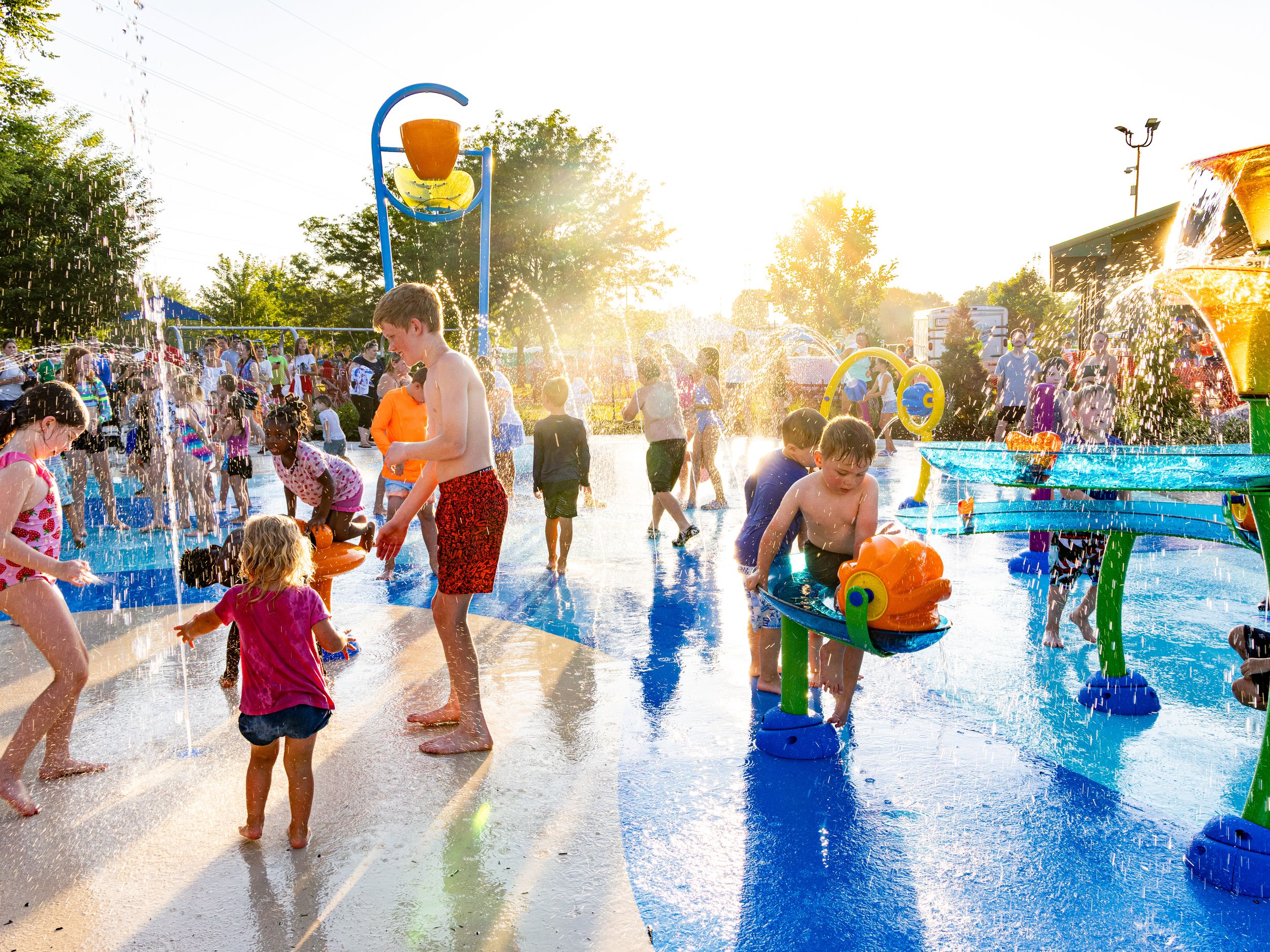 Children playing at a splash pad water park on a sunny day with water spraying and families enjoying summer fun.