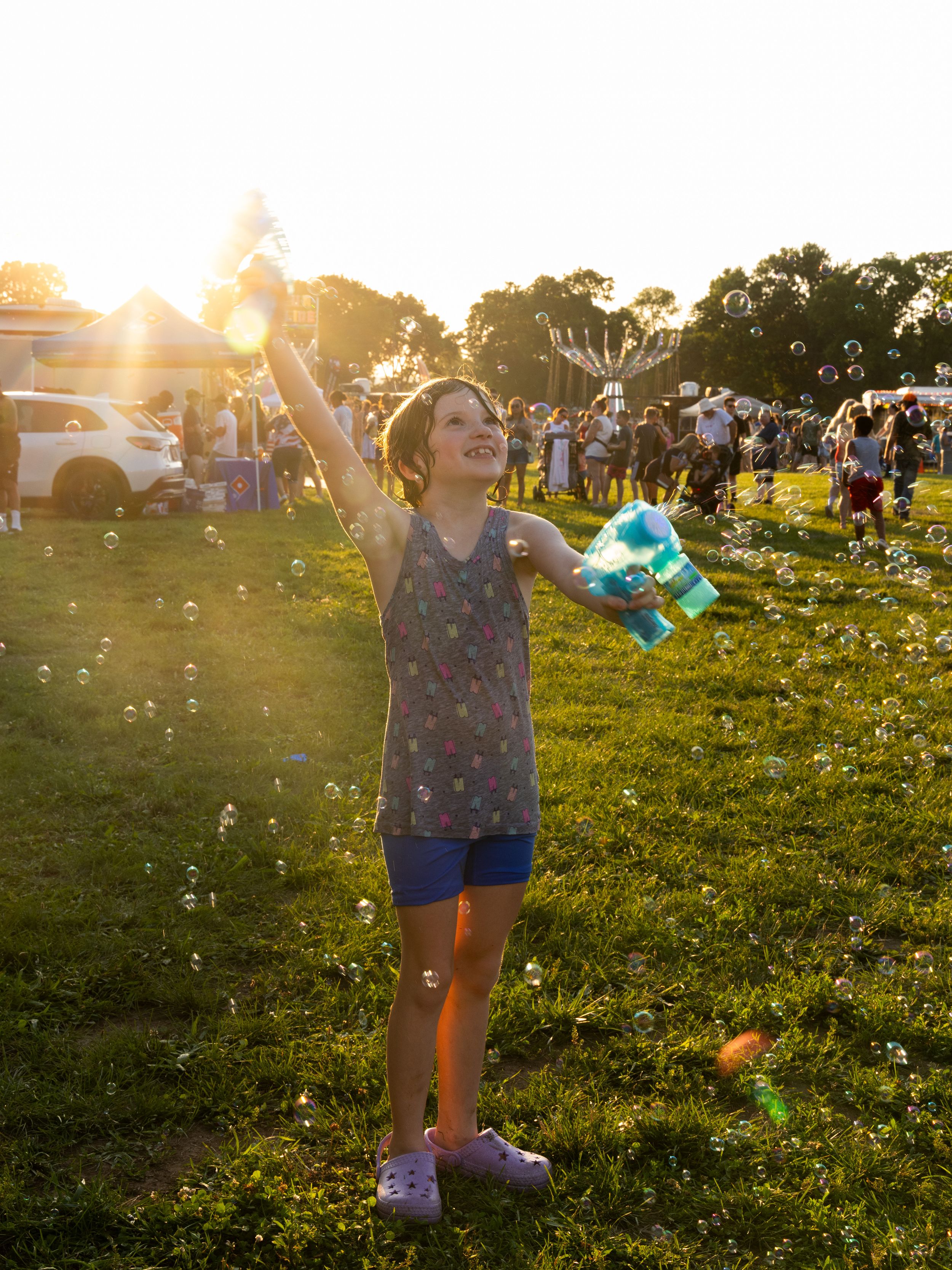 Young girl joyfully playing with bubbles at a sunny outdoor festival, raising her arm with a bubble machine in hand.