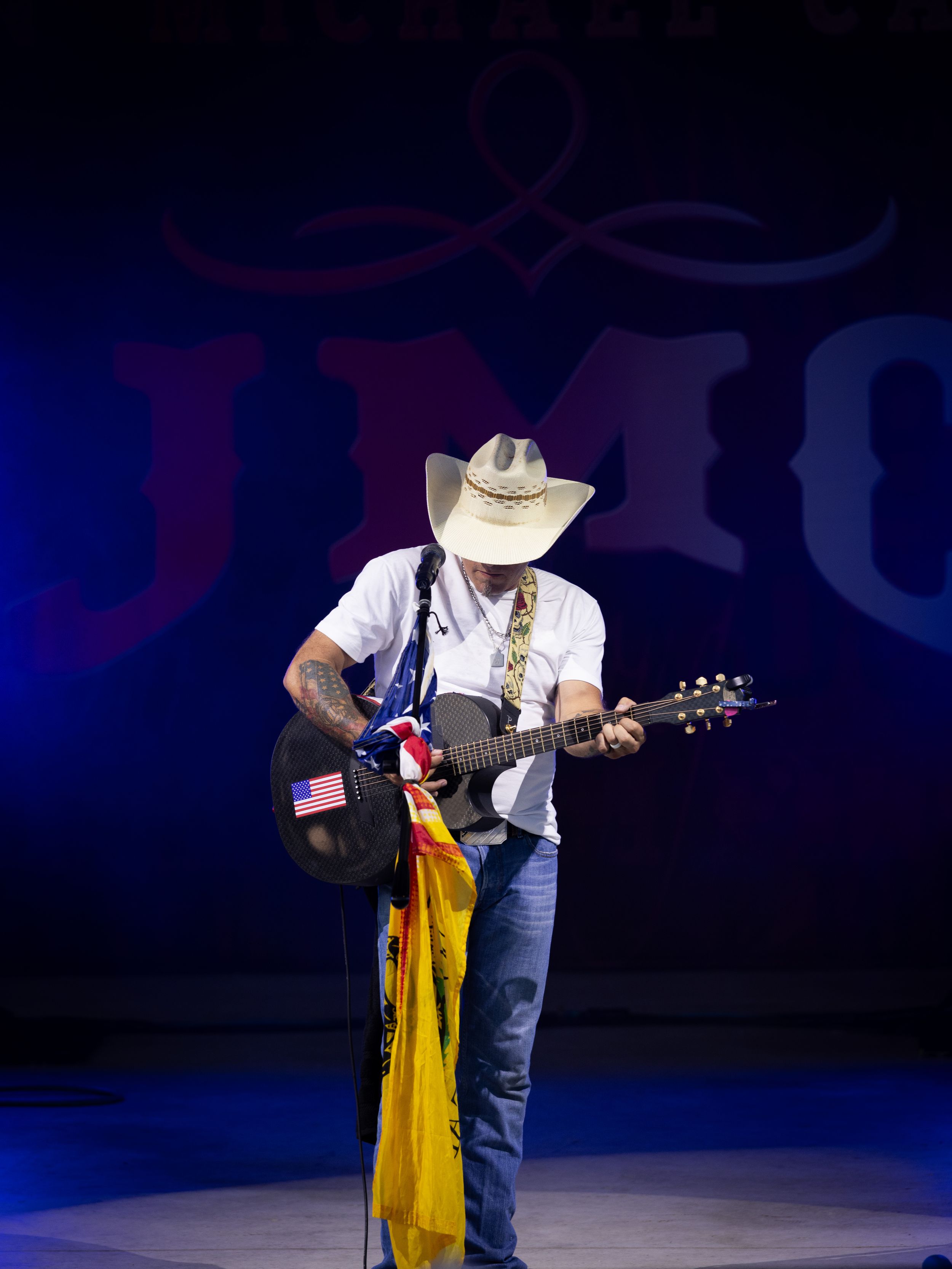 Country musician in cowboy hat plays guitar on stage with American flag, performing live at a concert event.