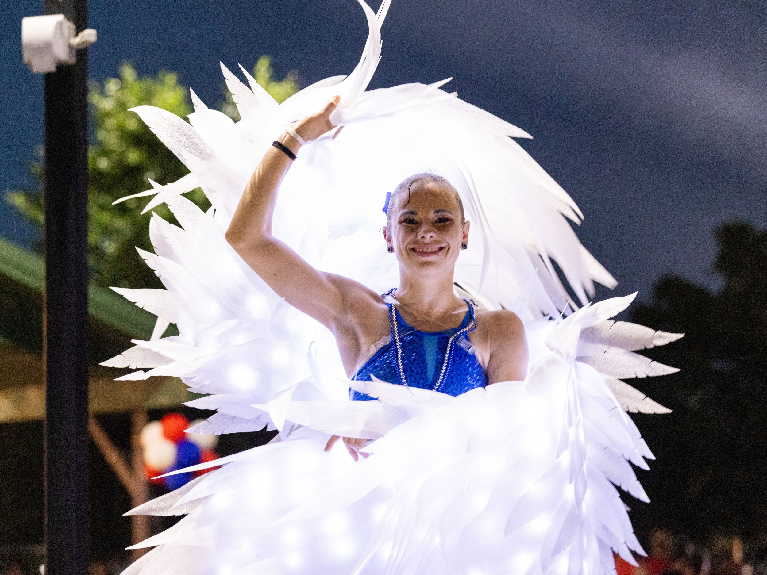 Performer in white feathered costume and blue outfit waves at a vibrant nighttime parade celebration.
